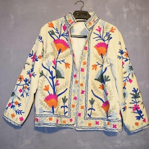 Peut inclure: Veste crème avec broderies florales vibrantes. La veste présente un col mandarin et est ornée de fleurs brodées colorées dans les tons de rose, orange, bleu et vert. La broderie est détaillée et couvre le devant, les manches et le col.