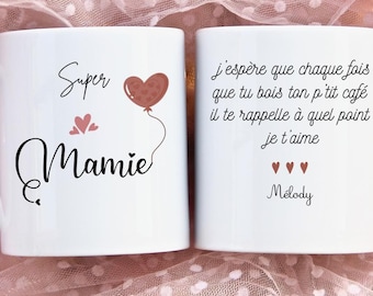 May include: Two white ceramic mugs with black text and pink hearts. The mug on the left says "Super Mamie" with a pink heart balloon. The mug on the right says "j'espère que chaque fois que tu bois ton p'tit café il te rappelle à quel point je t'aime" with three pink hearts and "Melody".
