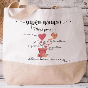 Peut inclure: Un sac fourre-tout en toile avec un fond et des poignées beiges. Le sac porte le texte "super nounou" et d'autres textes français avec une illustration de cœur et d'arrosoir. Un chapeau de paille est visible en arrière-plan.