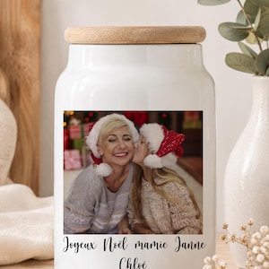 Peut inclure: Un bocal en céramique blanche avec un couvercle en bambou, orné d'une impression photographique carrée montrant un adulte et un enfant portant des bonnets de Noël, avec le texte Joyeux Noël mamie Janne Chloé écrit en lettres cursives noires.