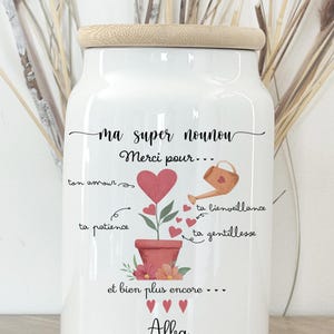 Peut inclure: Pot en céramique blanc avec couvercle en bois, orné de texte manuscrit et d'illustrations. Le pot présente un motif en forme de cœur avec les mots "ma super nounou" et "Merci pour..." écrits dessus. Le nom "Alba" est également écrit.