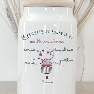 Könnte beinhalten: Weißes Keramikglas mit Holzdeckel, mit handschriftlichem Design auf Französisch. Der Text enthält "La recette du bonheur de ma Nounou d'amour" und eine Cupcake-Illustration. Das Glas ist ein charmantes Küchen-Dekorationselement.