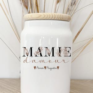 Peut inclure: Un pot en céramique blanc avec un couvercle en bois. Le pot porte l'inscription "MAMIE d'amour" en noir et marron, avec des motifs floraux. Les noms "Aimée Augustin" sont en dessous, avec des cœurs.