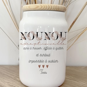 Puede incluir: Tarro de cerámica blanco con tapa de madera. El tarro tiene la palabra "NOUNOU" en escritura negra con detalles florales, y texto adicional en francés. El tarro está decorado con tres símbolos de corazón y el nombre "Tess".