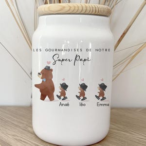 Könnte beinhalten: Weißes Keramikglas mit Holzdeckel, mit einer Bärenillustration mit drei kleineren Bärenfiguren. Der Text "LES GOURMANDISES DE NOTRE Super Papi" und die Namen Anaé, Léo und Emma sind auf dem Glas aufgedruckt.