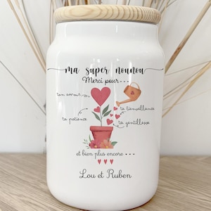 Peut inclure: Un bocal en céramique blanc avec un couvercle en bois, orné d'un motif floral et de texte. Le bocal porte l'inscription "ma super nounou Merci pour..." et les noms "Lou et Ruben".