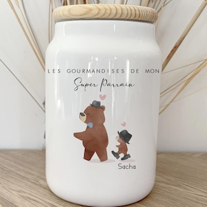 Peut inclure: Pot en céramique blanc avec un couvercle en bois. Le pot présente une illustration de dessin animé d'un ours et d'un enfant, tous deux portant des chapeaux. Le texte sur le pot se lit "LES GOURMANDISES DE MON Super Parrain" et le nom "Sacha".