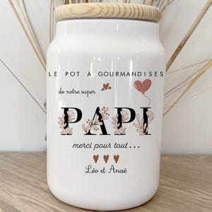 Könnte beinhalten: Ein weißes Keramikglas mit Holzdeckel, mit dem Text "LE POT À GOURMANDISES" und "PAPI" in dekorativer Schrift. Das Glas enthält den Text "merci pour tout..." und die Namen "Léo et Anaé".