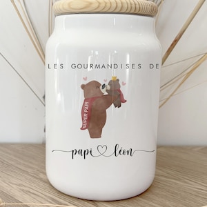 Puede incluir: Tarro de cerámica blanca con tapa de madera. El tarro presenta un diseño de oso de dibujos animados con el texto "LES GOURMANDISES DE papi ️ léon". El oso sostiene un oso más pequeño con corona y bufanda roja con "SUPER PAPI".