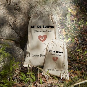 May include: Two beige drawstring bags with the text "KIT DE SURVIE Pour la maternite" and a heart-shaped balloon with footprints. The larger bag also has the text "Courage Papa". The smaller bag has the text "Courage Papa Jarvice".