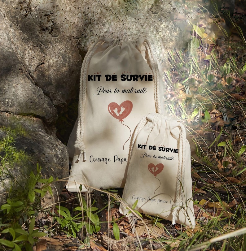 May include: Two beige drawstring bags with the text "KIT DE SURVIE Pour la maternite" and a heart-shaped balloon with footprints. The larger bag also has the text "Courage Papa". The smaller bag has the text "Courage Papa Jarvice".