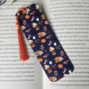 May include: A blue bookmark with a fall-themed pattern. The pattern includes pumpkins, mushrooms, cinnamon sticks, and a cup of hot chocolate with whipped cream. The text "Spice" is on the cup of hot chocolate.