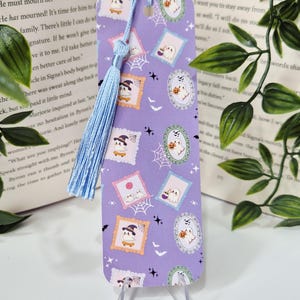 May include: A lavender bookmark with a repeating pattern of Halloween stamps, including ghosts, pumpkins, and spiderwebs. It has a light blue tassel. The bookmark is displayed on a clear stand.
