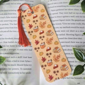 May include: A bookmark with an autumnal theme. It features a light orange background with illustrations of pumpkins, leaves, pie, coffee cups, and books. The bookmark is decorated with a red tassel.
