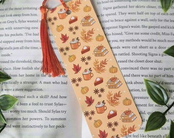 May include: A bookmark with a fall theme. It features a light orange background with illustrations of pumpkins, leaves, pie, coffee cups, and books. The bookmark is decorated with a red tassel.