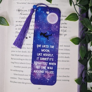 May include: A purple bookmark with a galaxy background and a silhouette of a child swinging on a swing. The bookmark has the text "She liked the moon, like herself. It shined it's brightest when no one was around to see."
