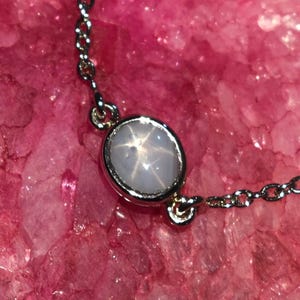 May include: A silver chain bracelet with an oval, pale blue star sapphire gemstone. The gemstone is set in a silver bezel and shows a six-rayed star pattern. The bracelet is displayed on a pink, crystalline surface.