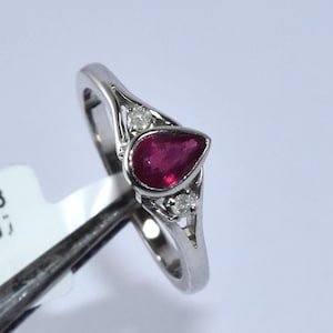 Op de afbeelding: Een ring met een peervormige robijn, gezet in witgoud, met twee kleinere diamanten aan weerszijden van de robijn. De ring ligt op een witte achtergrond.