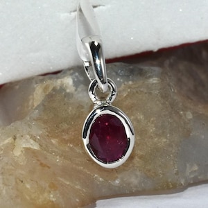 May include: A silver pendant with a deep red, oval-shaped gemstone. The gemstone is set in a silver bezel and hangs from a delicate silver chain. The pendant is displayed on a light brown rock against a white background.