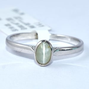 以下が含まれることがあります： 楕円形の淡い緑色の宝石が付いたシルバーリング。リングはシンプルなバンドと宝石用のベゼルセッティングを備えています。リングは白い背景に表示されています。