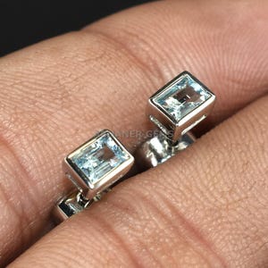 Può includere: Un paio di orecchini a lobo in argento, ciascuno con una pietra preziosa rettangolare azzurra. Gli orecchini sono posti su uno sfondo scuro, evidenziando la brillantezza delle pietre preziose. Piccoli e delicati, adatti all'uso quotidiano.