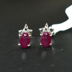 May include: A pair of ruby stud earrings. Each earring features an oval, deep red ruby gemstone set in a silver metal setting. The setting has a decorative design at the top. The earrings are displayed on a dark surface.