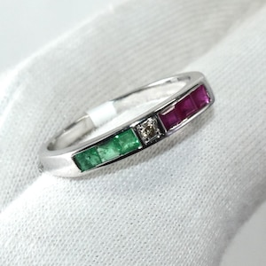 Peut inclure: Une bague en or blanc avec trois pierres précieuses de forme carrée. La bague présente deux émeraudes vertes et un rubis rouge, avec un petit diamant entre les émeraudes et le rubis.