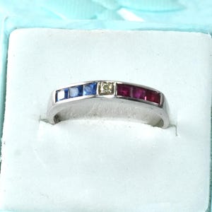 May include: A silver ring with a row of blue, yellow, and red gemstones. The ring is displayed in a light blue gift box with a matching ribbon. The gemstones are rectangular and set in a silver band.
