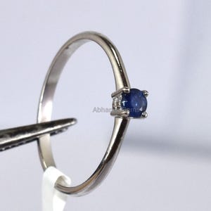 Pode incluir: Um delicado anel de prata com uma pequena pedra preciosa redonda azul escuro. O anel é segurado por uma pinça, com uma etiqueta branca anexada. A etiqueta tem o número "0,23" impresso. O anel está em um fundo branco.