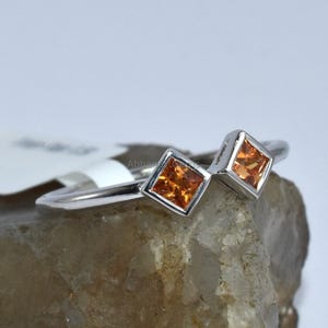 May include: A silver ring with two square-shaped orange gemstones. The ring is set on a rock.
