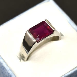 Könnte beinhalten: Ein silberner Ring mit einem rechteckigen, tiefroten Edelstein. Der Ring hat ein schlichtes, modernes Design und wird auf einer weißen Oberfläche in einer hellblauen Box präsentiert. Der Edelstein ist der Mittelpunkt und unterstreicht seine satte Farbe und seinen Schliff.