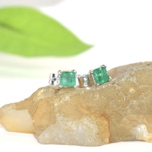 May include: A pair of stud earrings featuring square-cut green emerald gemstones set in silver metal. The earrings are positioned on a textured, light brown, irregular stone surface with a blurred green leaf visible in the background.
