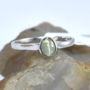 Puede incluir: Un anillo de plata con una piedra ovalada verde claro. El anillo está colocado sobre una roca.