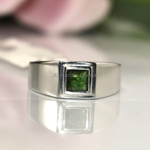 Könnte beinhalten: Ein silberner Ring mit einem quadratischen, smaragdgrünen Edelstein in der Mitte. Der Ring hat ein schlichtes, modernes Design mit poliertem Finish. Der Edelstein hat eine leuchtend grüne Farbe, und der Ring wird auf einer reflektierenden Oberfläche präsentiert.