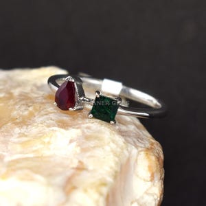 May include: A silver ring with a pear-shaped red gemstone and a square green gemstone. The ring is displayed against a neutral backdrop, accentuating the contrast between the gemstones and the metal.