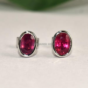 May include: A pair of oval-shaped ruby stud earrings. Each earring features a vibrant red gemstone set in a silver-colored bezel. The earrings are displayed on a white surface, with a green leaf and a branch in the background.