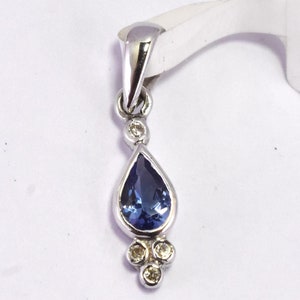 Op de afbeelding: Zilveren hanger met een peervormige, donkerblauwe edelsteen en drie kleinere, ronde, heldere edelstenen. De hanger hangt aan een zilveren balein. Gepresenteerd tegen een witte achtergrond.