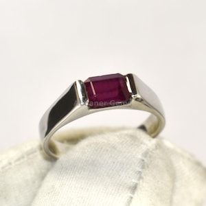 May include: A silver ring with a rectangular, deep red gemstone. The ring has a simple, modern design with a polished finish. The gemstone is set in a bezel setting, and the ring is held by a gloved hand.