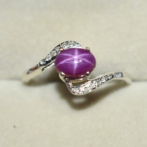 May include: A silver ring with a large, oval, star ruby gemstone. The ring has a swirling design with small, clear accent stones. The ring is displayed in a maroon jewelry box with a cream-colored interior.