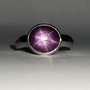 Puede incluir: Un anillo de plata con una gran gema de rubí estrella redonda. El rubí es de color rojo púrpura intenso con un patrón de estrella de seis rayos. El anillo tiene una banda simple y pulida.