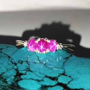 Puede incluir: Un anillo de plata con tres piedras preciosas rosas vibrantes de forma ovalada. El anillo está engastado con pequeñas piedras de acento transparentes a ambos lados de las gemas centrales. El anillo se muestra sobre una superficie turquesa texturizada.