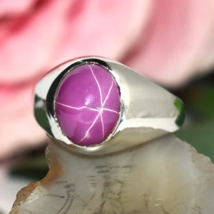 Könnte beinhalten: Ein silberner Ring mit einem großen, ovalen, purpurrosa Sternrubin-Edelstein. Der Edelstein weist ein sechsstrahliges Sternmuster auf. Der Ring ist auf einer natürlichen, mehrfarbigen Steinoberfläche platziert, mit einer verschwommenen rosa Blume im Hintergrund.