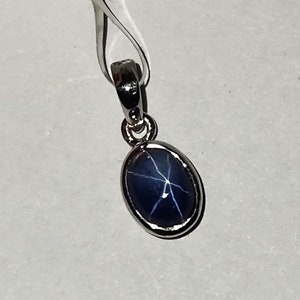 Könnte beinhalten: Silberanhänger mit einem ovalen, dunkelblauen Edelstein mit sternförmigem Muster. Der Edelstein ist in eine silberne Lünette gefasst und an einer silbernen Öse aufgehängt. Der Anhänger wird vor weißem Hintergrund gezeigt.