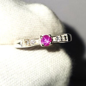 Puede incluir: Un anillo de plata con una gema rosa central en forma de estrella. El anillo está adornado con pequeñas gemas claras a cada lado de la piedra central. El anillo se muestra sobre una superficie blanca.