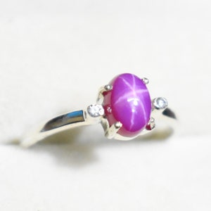 Può includere: Un anello d'argento con una vibrante gemma di rubino a stella taglio ovale. Il rubino è di un colore rosa intenso, con un effetto stella a sei raggi. Due piccole gemme rotonde e trasparenti affiancano il rubino. L'anello è in un cofanetto.