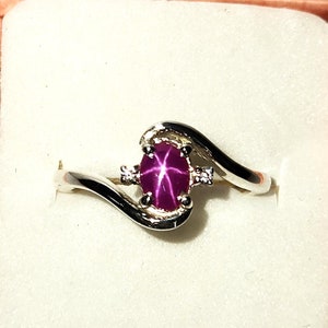 Peut inclure: Une bague en argent avec une pierre précieuse en rubis étoilé rose et deux diamants plus petits. La bague est dans une boîte blanche avec un couvercle rose.