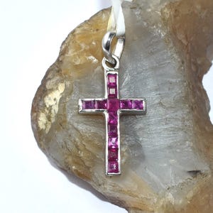 Puede incluir: Un colgante de cruz plateada con piedras preciosas cuadradas de color magenta. La cruz se muestra sobre una superficie clara y rugosa. Una pequeña etiqueta está adherida al colgante.