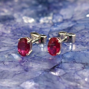 May include: Silver stud earrings, each featuring an oval-cut, deep red gemstone. The earrings are presented on a blue and white marbled surface. The design highlights the gemstones' rich colour and the jewellery's understated style.