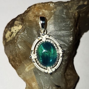 Puede incluir: Un colgante de plata con una piedra preciosa ovalada de color verde azulado. La piedra preciosa está engastada en un marco de plata decorativo con un borde detallado. El colgante se muestra sobre una piedra gris rugosa sobre un fondo blanco. Es una joya.
