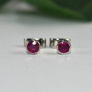 May include: A pair of small stud earrings. Each earring has a vibrant, round, ruby-coloured gemstone set in a silver metal bezel. The earrings are displayed on a white surface with a blurred green background.